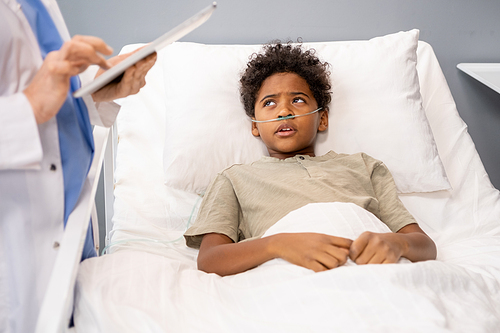 African little boy lying in bed and listening to recommendations of doctor who visiting him in hospital ward