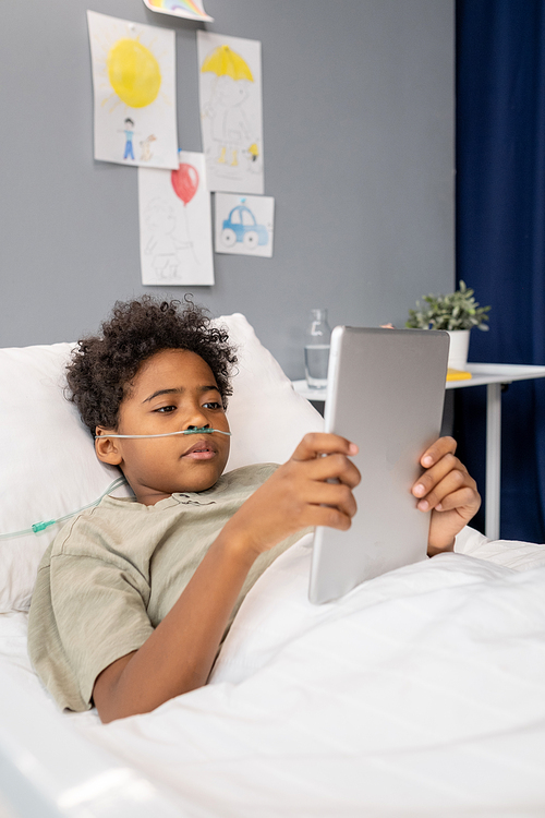 African little boy watching something on digital tablet while lying on bed in hospital ward