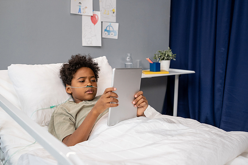 African little boy reading online on digital tablet while lying in bed at hospital ward