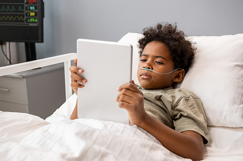 Sick little boy lying in bed and reading online book on digital tablet during his treating at hospital