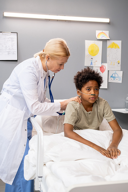 Mature female doctor in white coat examining the little boy with stethoscope while he sitting in bed at hospital ward, she listening to his breathing