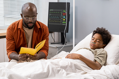 African father reading a book to his little son while he sleeping in hospital bed