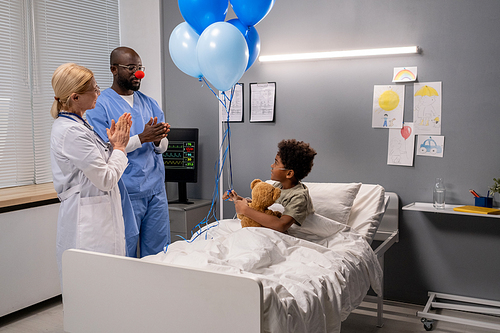 Two doctors clapping hands and having fun the little boy while he sitting in bed with toy and balloons, they congratulating him with recovering