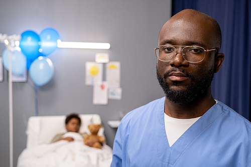 Portrait of African male doctor in eyeglasses and uniform looking at camera while working in child department at hospital