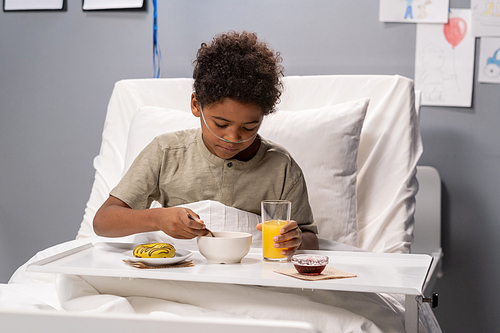 Sick little boy sitting in bed with tray and having breakfast while being treated at hospital