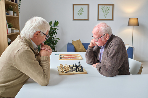 Two senior men sitting at the table and playing chess together in the room