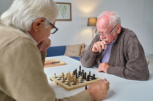 Senior man in eyeglasses thinking over his step while playing chess together with friend in the nursing home