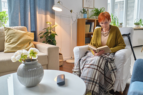 Senior woman sitting in armchair under the blanket and reading a book in the living room
