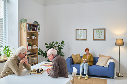 Two senior men sitting at the table and playing chess with woman reading a book on the sofa, they spending time in nursing home