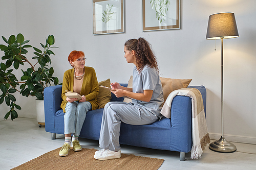 Young nurse giving recommendations to senior woman while they sitting on sofa in the room