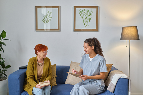 Elderly woman discussing with nurse her treatment while they sitting on sofa in the room