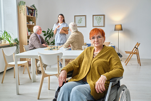 Portrait of mature woman in eyeglasses using wheelchair looking at camera sitting in the room with other senior people in the background