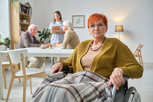 Portrait of elderly woman using wheelchair looking at camera, with nurse and other people in the background in the room
