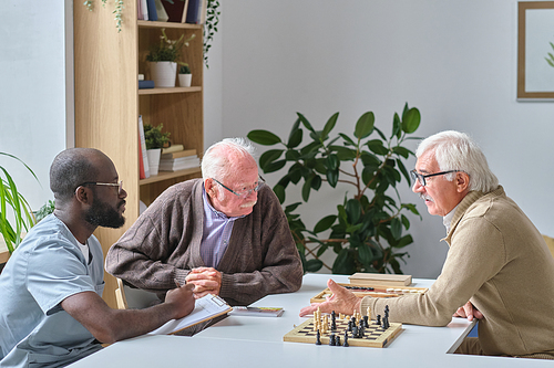 Senior man talking to African doctor who sitting together with two senior men while they playing chess in nursing home