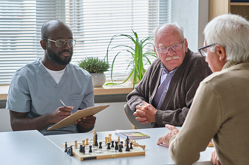 African doctor making notes in medical card while talking to elderly man during his chess game with friend at the table