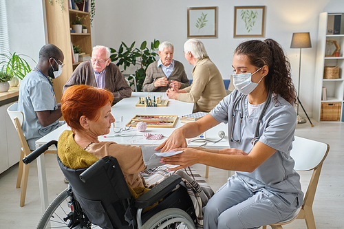 Nurse measuring the pressure to senior woman who using wheelchair in the nursing home with other senior people playing chess in the background