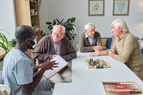Male doctor working in nursing home, he prescribing medicine to senior man while they sitting at the table with other men playing chess in the background