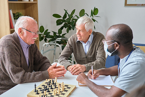Senior men sitting at the table with doctor and discussing their treatment during chess game