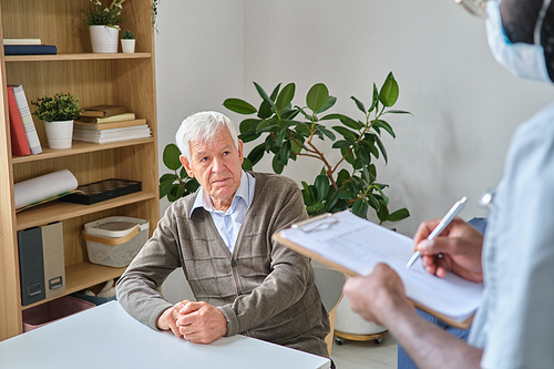 Elderly man sitting at the table and listening the prescription of his doctor while he making notes in medical card