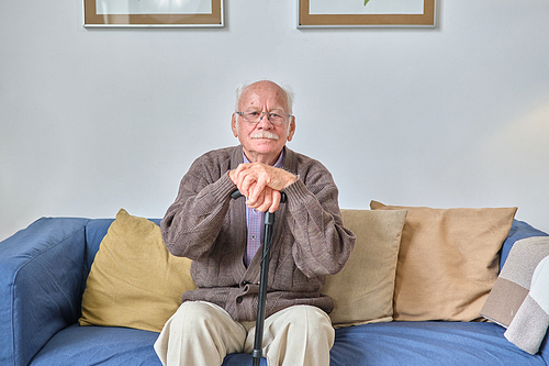 Portrait of senior man in eyeglasses sitting on sofa with crutch and looking at camera during his rest