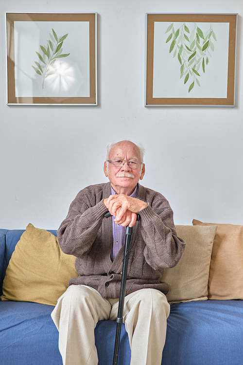 Portrait of senior man in eyeglasses looking at camera while sitting on sofa with crutch