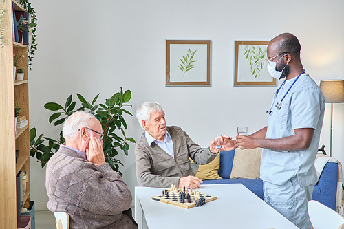 Male doctor giving the glass of water and pills to senior man while he sitting at the table and playing chess with his friend