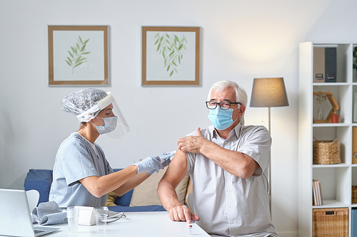 Elderly man in mask getting vaccine in his shoulder by the nurse in uniform while sitting at doctors office