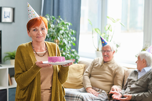 Portrait of senior woman in party hat looking at camera holding birthday cake with candle while standing in the room with her friends in the background