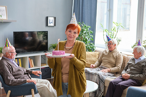 Portrait of elderly woman in hat holding birthday cake with candle and looking at camera while standing in the room with her friends