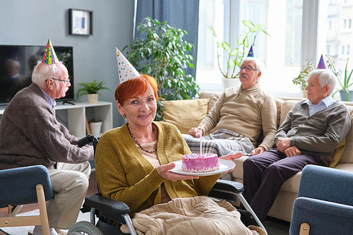 Portrait of elderly woman using wheelchair holding cake and smiling at camera while celebrating birthday with her friends