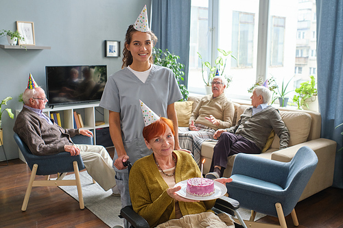 Portrait of senior woman sitting in wheelchair smiling at camera and holding a birthday cake with nurse standing behind her, they celebrating birthday in nursing home