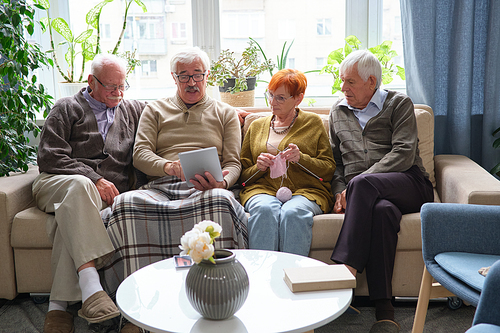 Senior man sitting on sofa together with his friends in the room and showing something on digital tablet