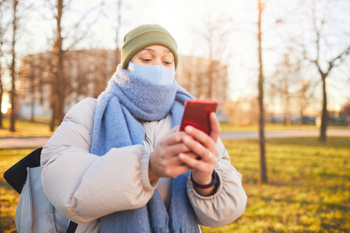 Woman in warm clothing and in mask using navigator on her mobile phone while walking in the city