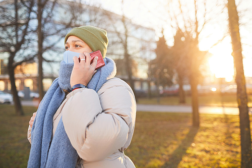 Young woman in protective mask talking on the phone while walking in the city