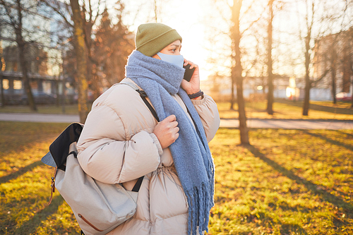 Woman in warm clothing and in mask has a conversation on her mobile phone while walking outdoors