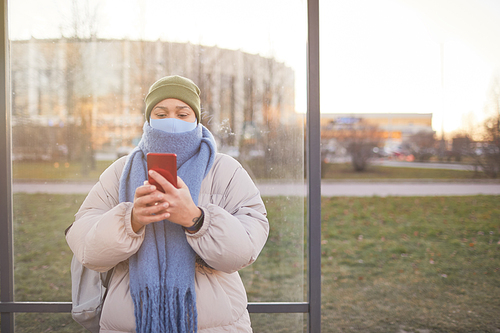 Young woman in protective mask and in warm clothing using her mobile phone while standing on bus stop