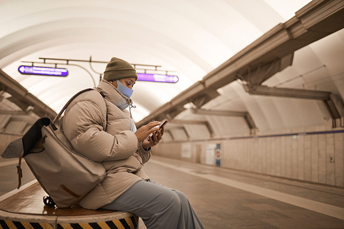 Young woman in warm clothing and in mask using her mobile phone while sitting underground