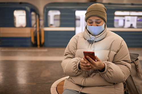 Young woman in mask and in warm clothing sitting in subway and using her mobile phone while waiting for her train