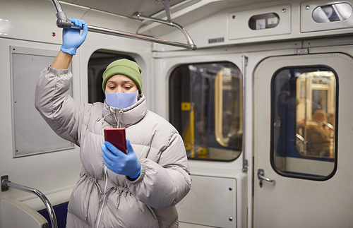 Woman in warm clothing and in protective mask and gloves reading online book on the phone while standing on a train