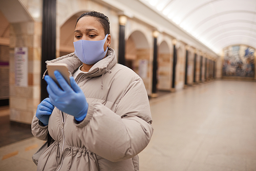 Blogger in mask and in warm clothing using her mobile phone while standing underground