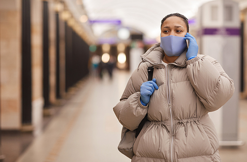 Young woman in warm clothing and in protective mask and gloves talking on the phone while walking along the underground