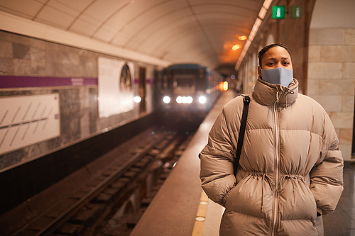 Portrait of young woman in protective mask looking at camera while standing in underground with train in the background