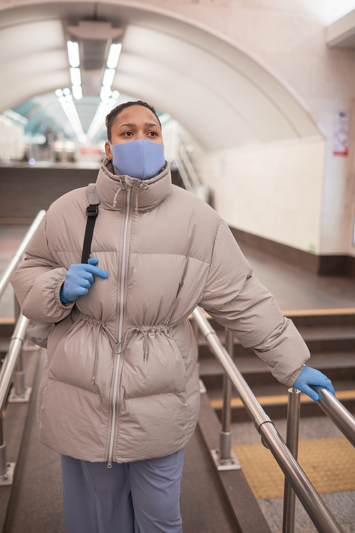 Young woman in protective mask and gloves standing on escalator in the underground