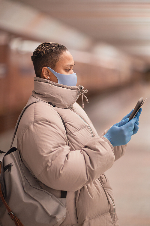 Side view of woman in protective mask standing and reading online book on her mobile phone