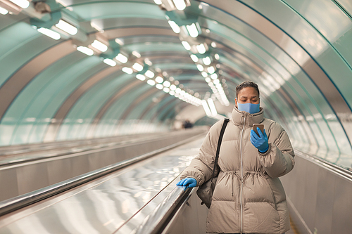 Young woman in mask standing on escalator and reading a message on her mobile phone