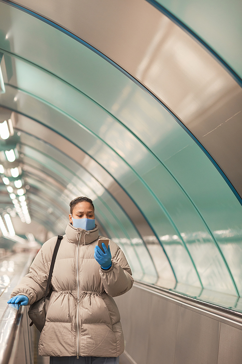 Young woman in protective mask and gloves using her mobile phone while standing on escalator