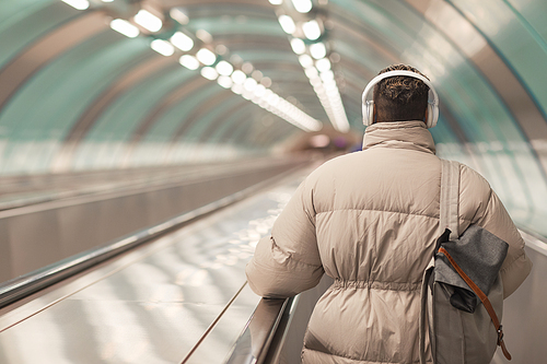 Rear view of plump woman in headphones with bag behind her back riding on escalator