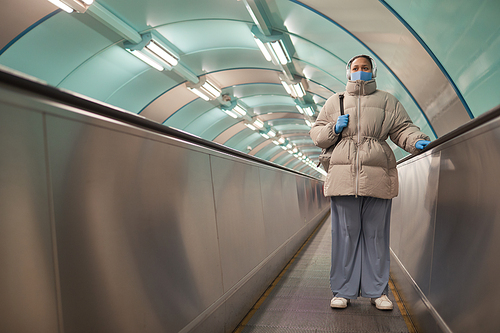 Plump woman in warm clothing wearing mask and gloves riding on escalator during pandemic