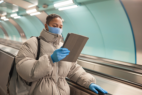 African woman in mask and in warm clothing reading a book on tablet pc while standing on escalator