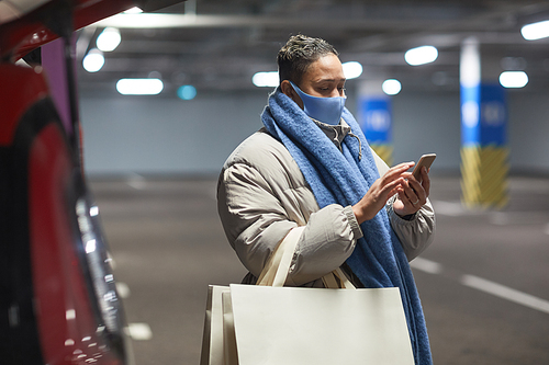 Young woman in protective mask with shopping bags using mobile phone while standing in an underground parking